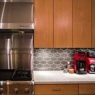 carbon gray stretch hex ceramic tile laid horizontally as a kitchen backsplash between a marble counter top and tall cabinets. This unique ceramic tile is a way to add fun, modern style to kitchens, bathrooms & features